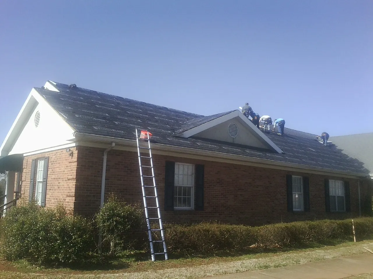 Crew tearing shingles off a brick ranch roof with a ladder set — Emergency Roof Repair work by our crew in Chestnut Hill
