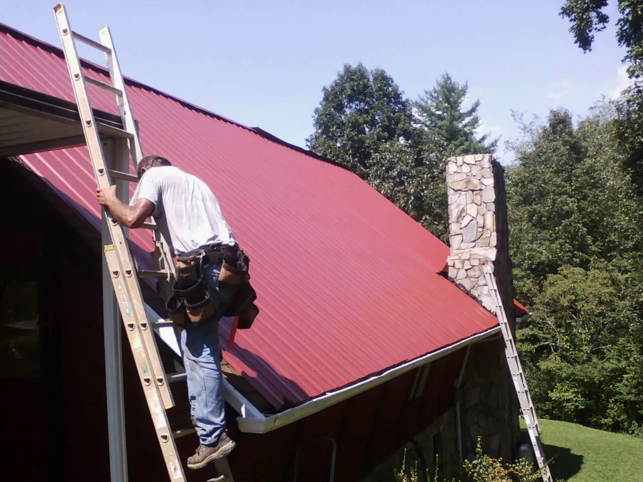 Roofer on a ladder beside a red metal roof and stone chimney — Soffit & Fascia Repair by Roofing Brothers in Chestnut Hill